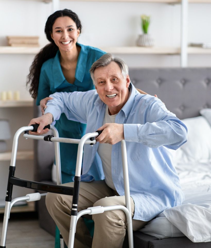 female-nurse-helping-elderly-male-with-walking-frame-stand-up-from-bed-at-home-professional-care.jpg